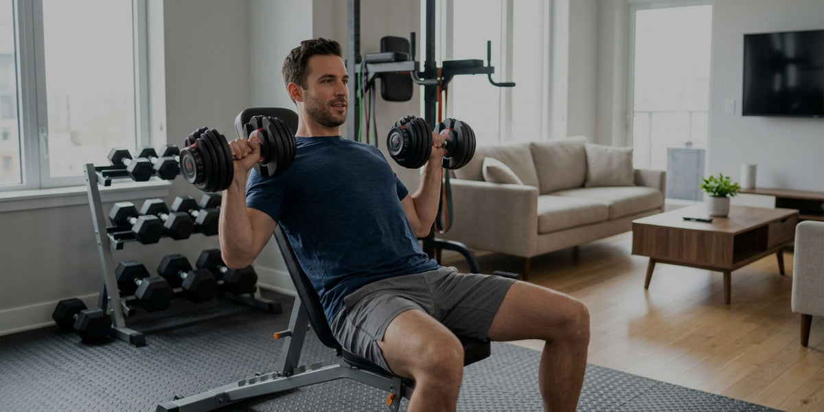 A man in a home gym setting, seated on an incline bench holding dumbbells, with a sofa and coffee table visible in the background.