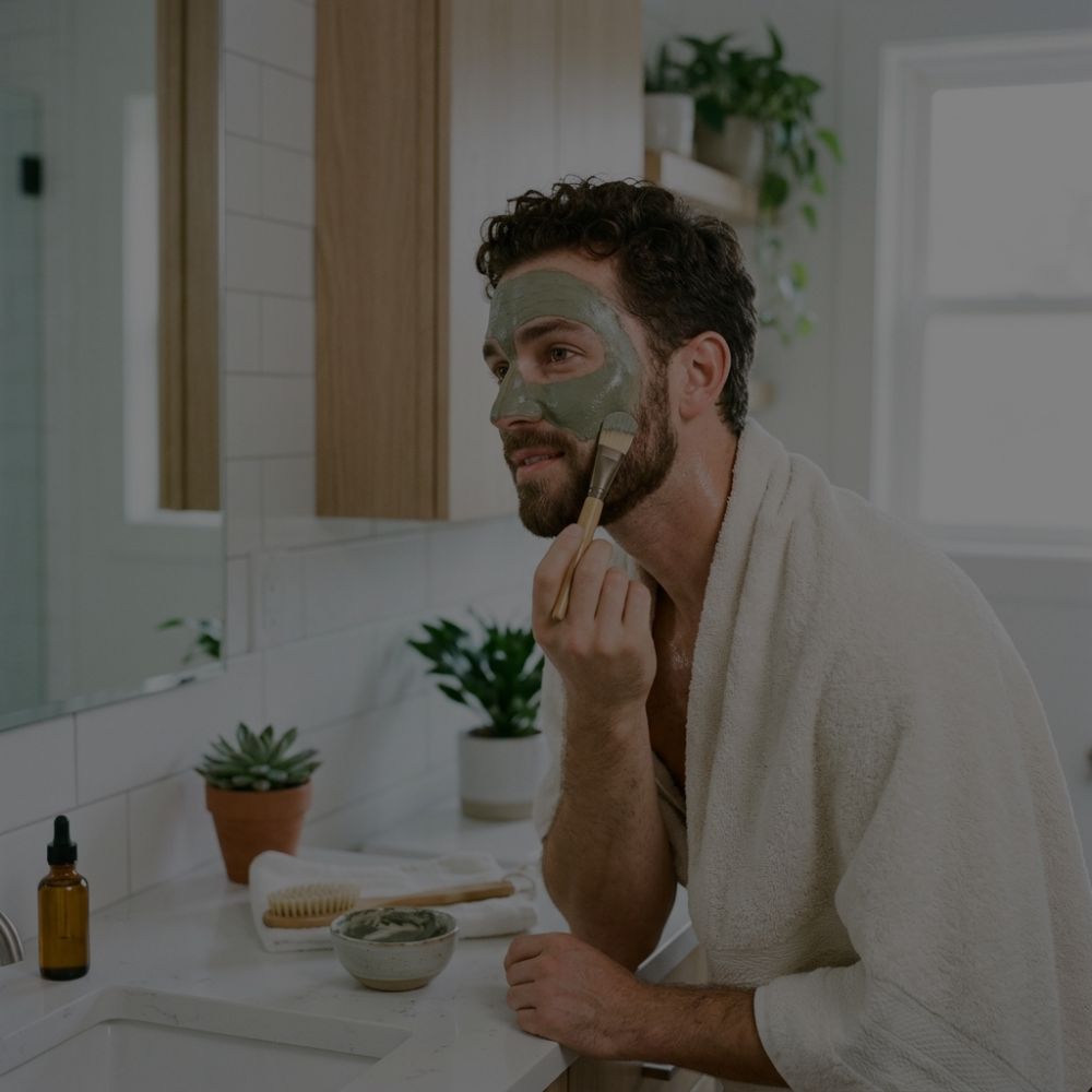 A man with a beard and a white towel around his shoulders applies a green face mask with a brush in a bright bathroom setting.